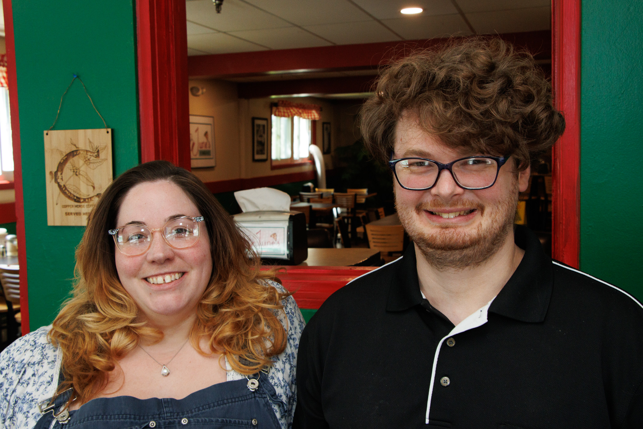 Two people smiling at the camera in a doorway with green walls and red trim, interior cafe-like space in the background.
