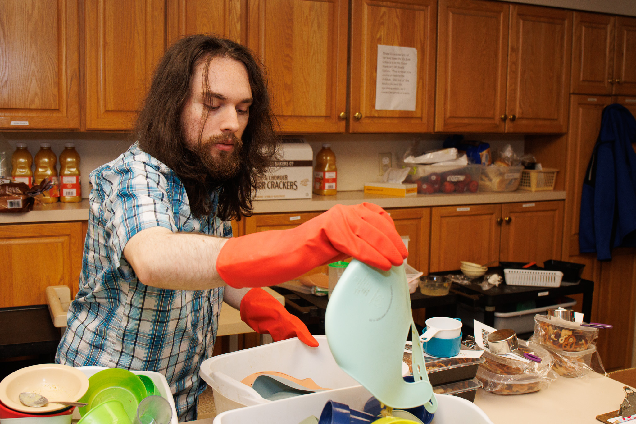 Man with long hair and a beard wearing an orange glove and a blue plaid shirt washes dishes in a cluttered kitchen.